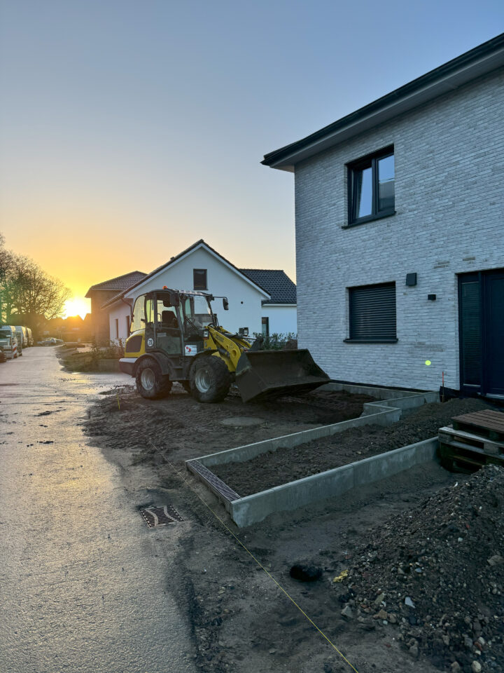 Ein zweistöckiges Haus mit Erdarbeiten im Vordergrund und einem Gehweg, der zur Tür führt, im Hintergrund, begleitet von einem Bagger.