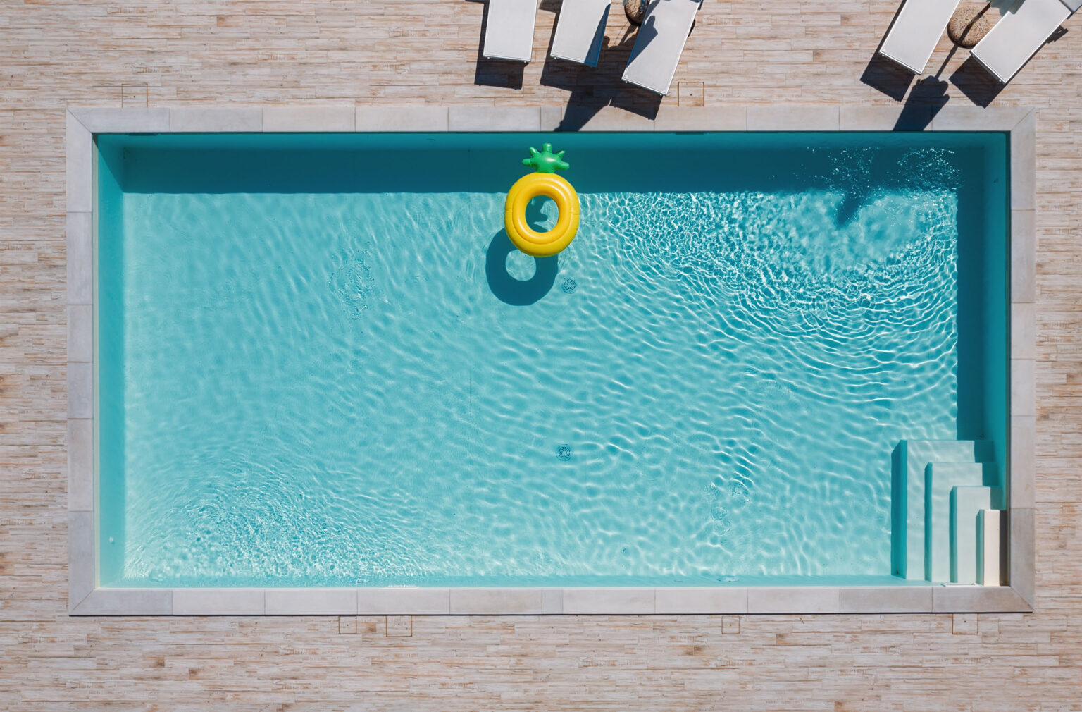 Rechteckiger blauer Swimmingpool mit Liegestühlen und Sonnenschirmen und großem aufblasbaren gelben Ananas-Schwimmschlauch.